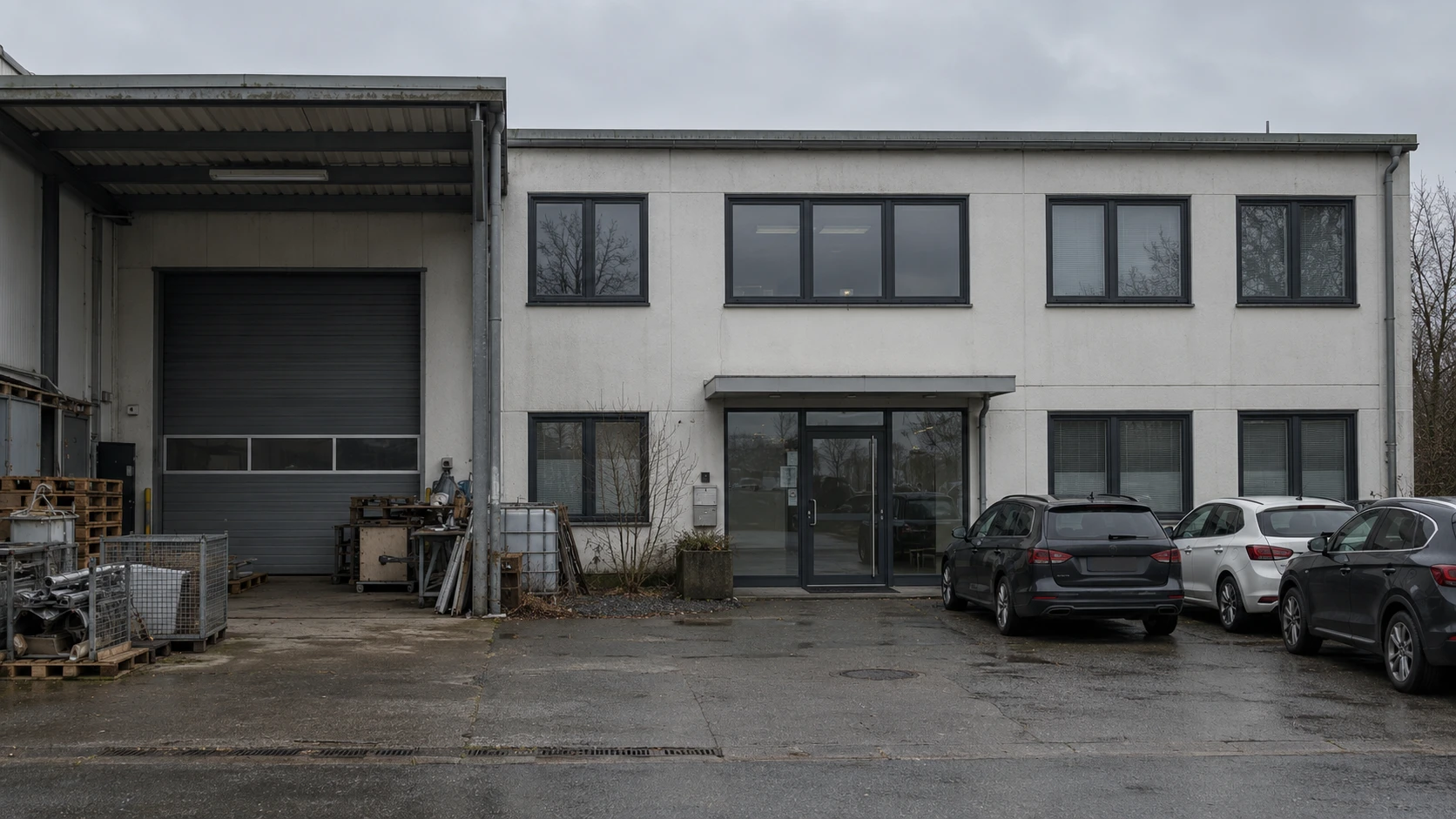 Vorher: Firmengebäude mit Rolltor bei grauem Himmel, davor Paletten, Gitterboxen und parkende Autos
