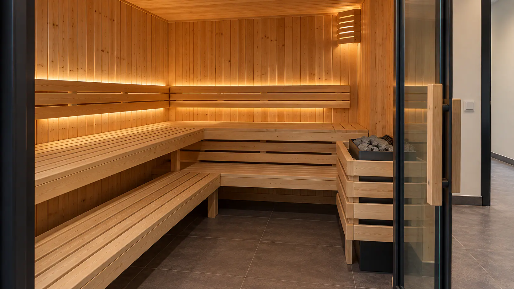Sauna mit hellen Holzpaneelen, zwei Bankhöhen und Ofen mit Schutzgitter.